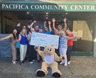 Pacifica Senior Services group waving outside the Pacifica Community Center with a Pacificans Care check and the bear mascot