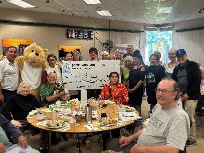 Seniors and the bear mascot gathered around a table of food at a Pacifica Senior Services lunch